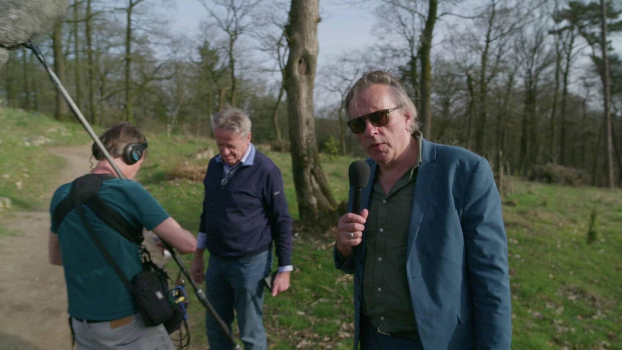 Man met een zonnebril staat met een microfoon in zijn hand in de natuur