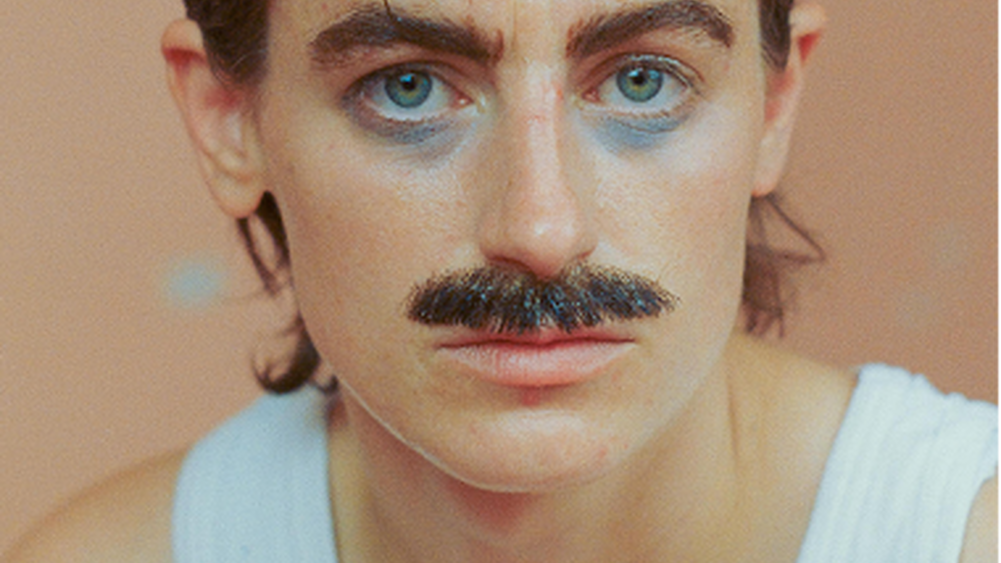 Een foto van Elmer. Ze zit tegen een licht oranje achtergrond en kijkt recht de camera in. Ze heeft een snor en een wit hemd aan.