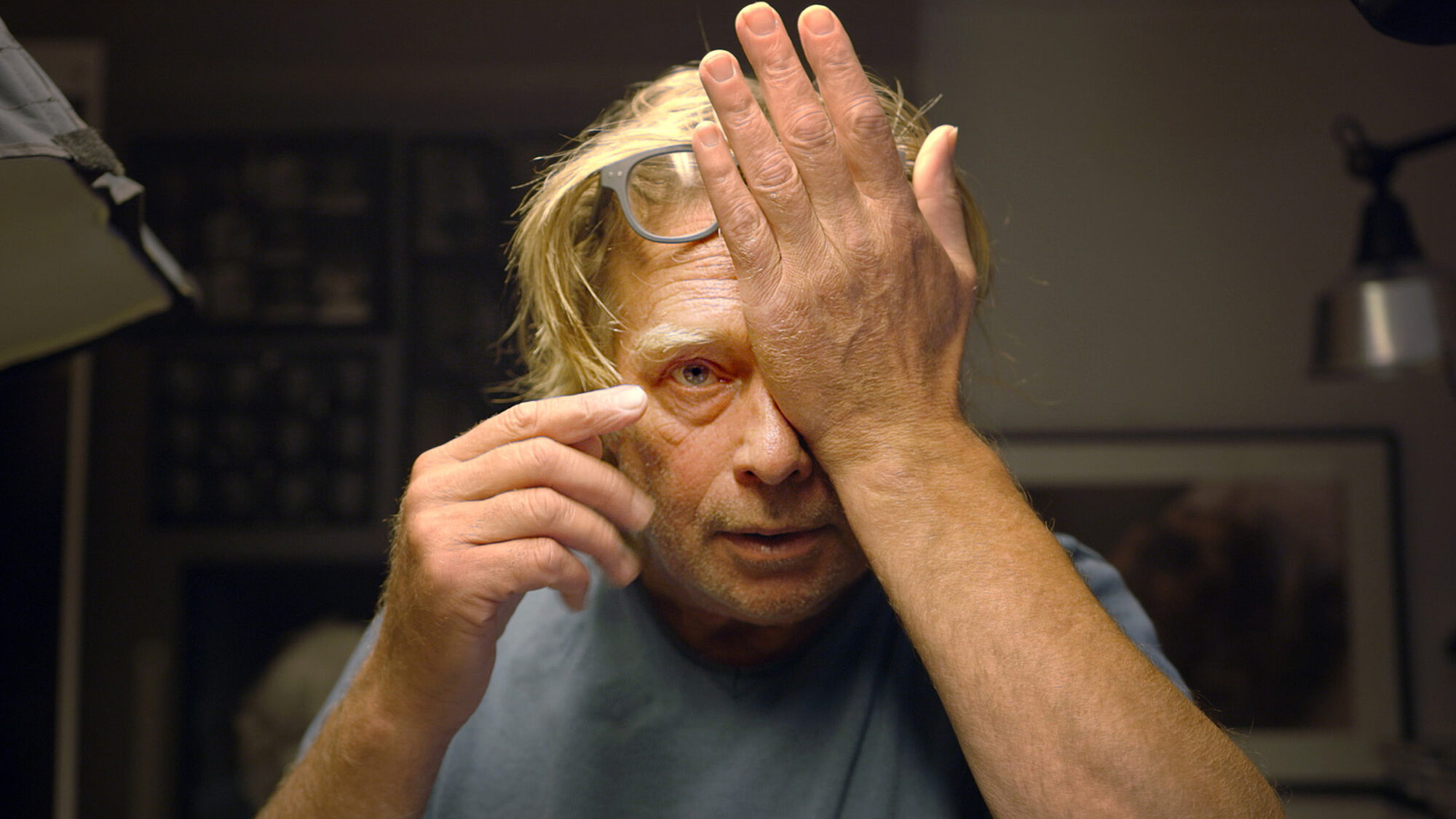 Een stil uit de documentaire. Je ziet fotograaf Koos Breukel recht de camera inkijken met een hand voor zijn linkeroog. Hij is goed uitgelicht en achter hem zie je een soort werkruimte.