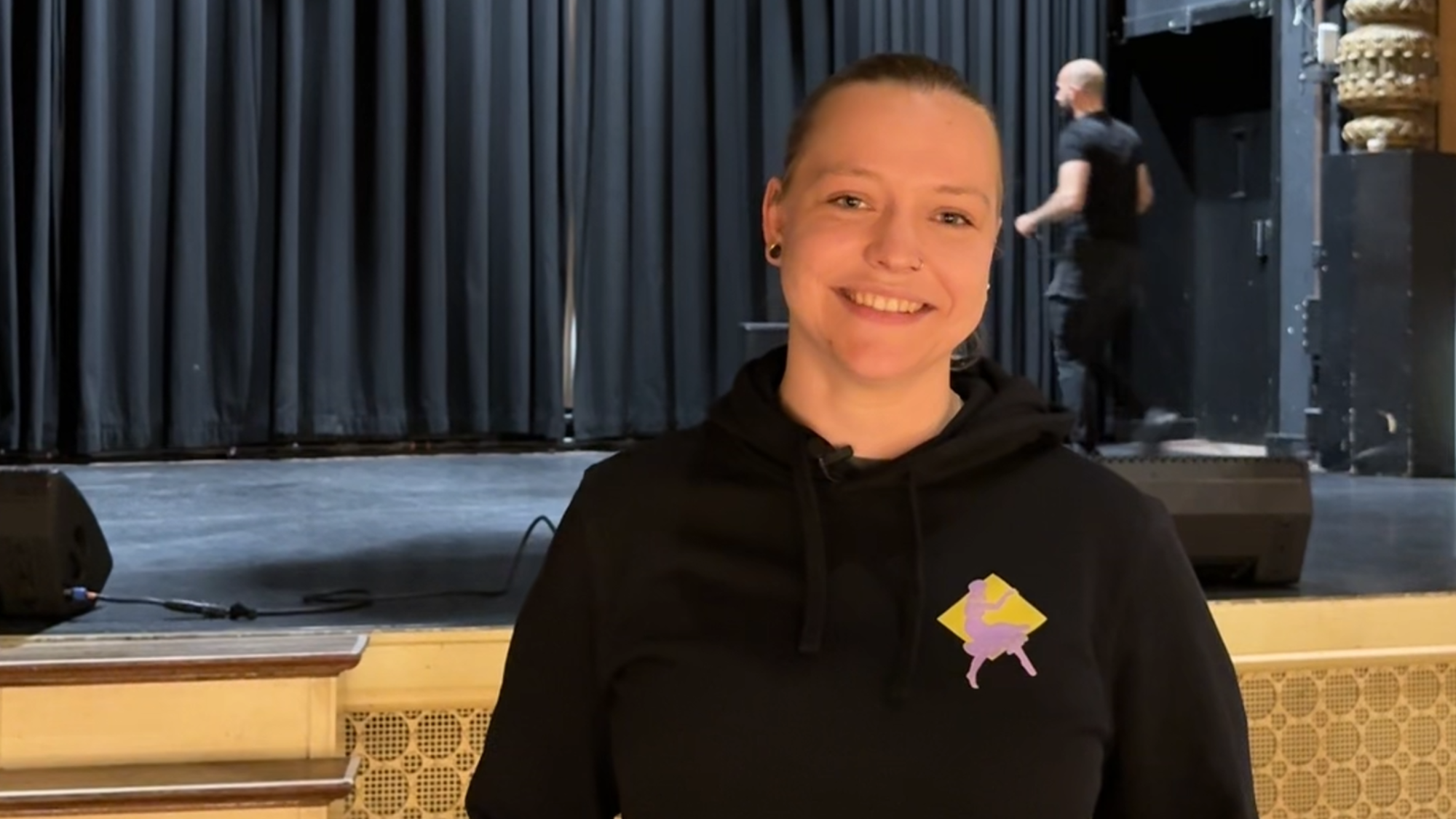 Izanka, de geïnterviewde, in de zaal. Ze heeft zwarte kleren aan en heeft haar handen in haar zakken. Ze kijkt lachend de camera in.