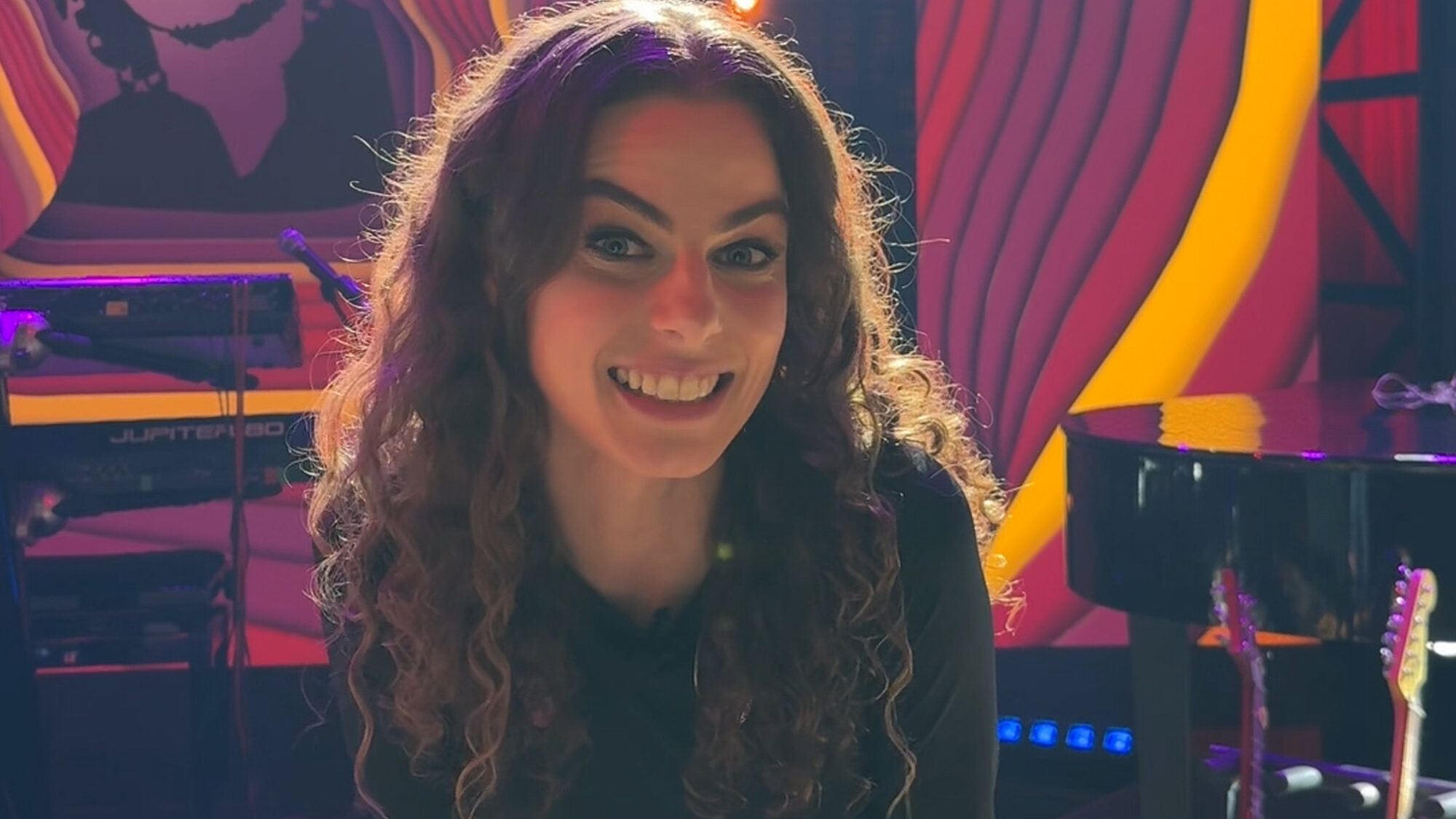 Yvette zit op een stoel in de tv studio van The Best of Pop en kijkt de camera in. Ze heeft lange bruine krullen en lacht.