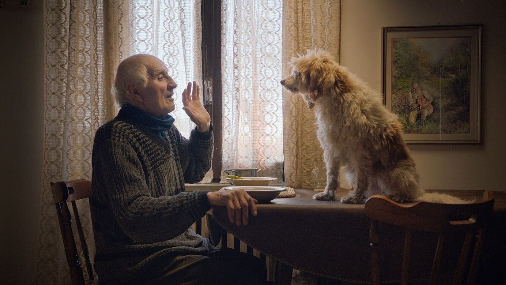 Man uit de documentaire zit aan tafel en praat tegen zijn hond die op de tafel zit.