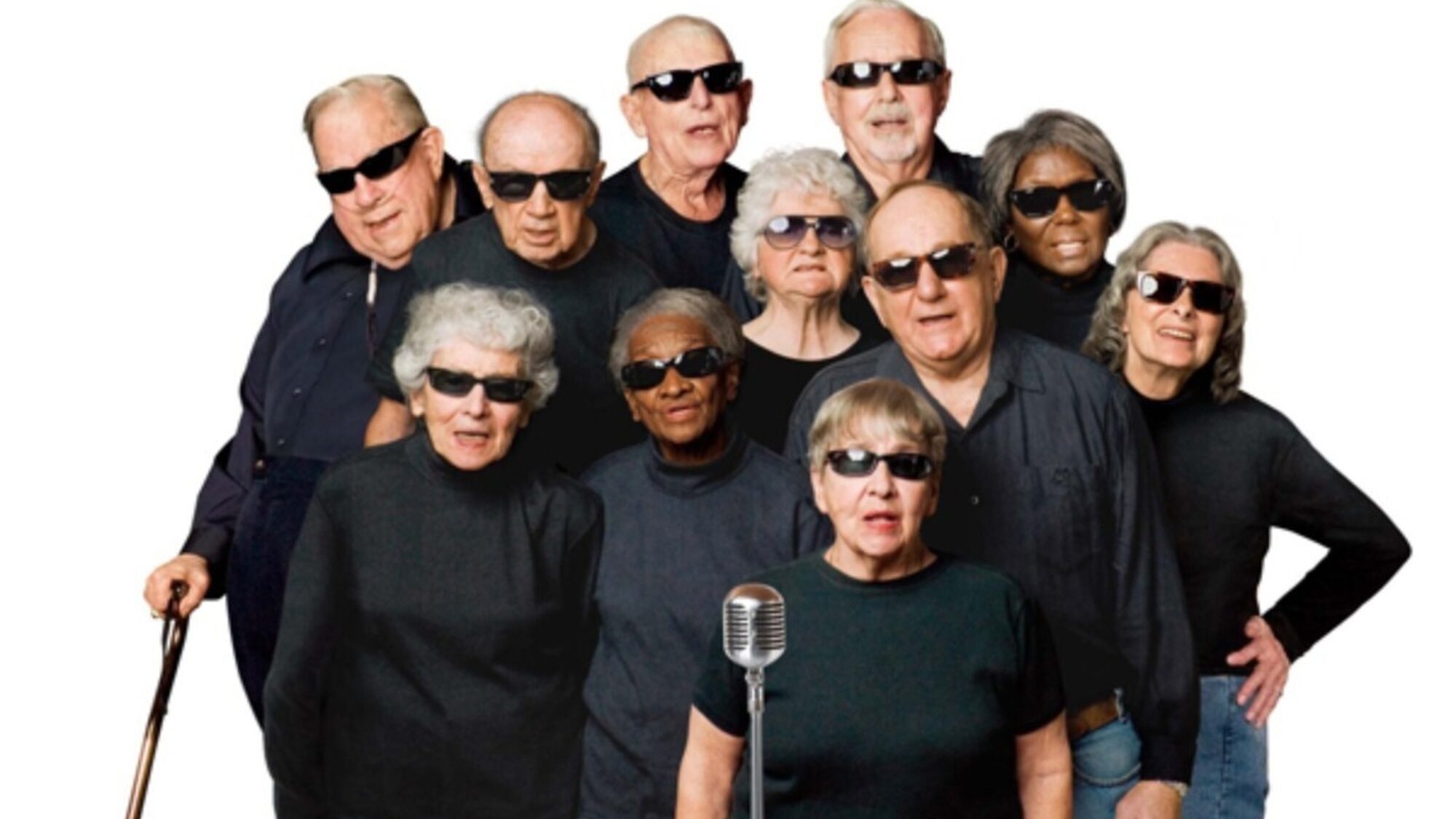 Een groep koorleden uit de documentaire zingt voor een microfoon. Ze dragen zwarte shirts en zwarte zonnebrillen.