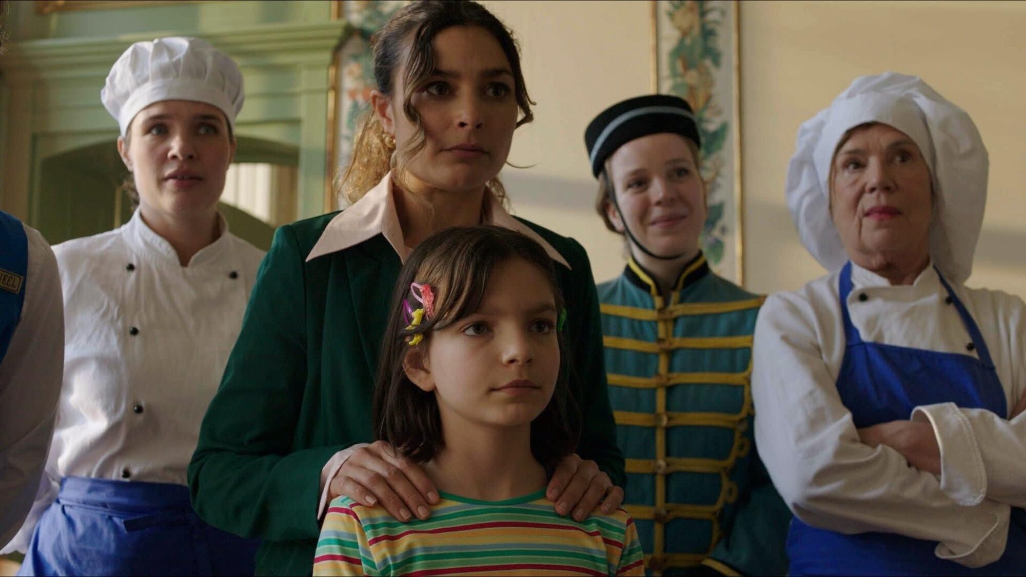 je ziet een moeder - hoteleigenaar met een stressvolle blik. 2 koks met witte en blauwe kleding, een portier met pet in een groene outfit en een klein meisje in een gestreepte trui