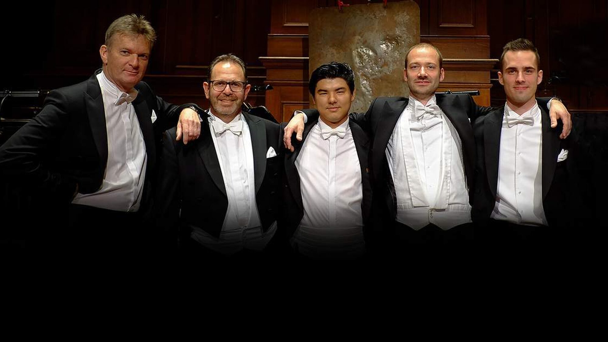 Een foto van de 5 paukenisten en slagwerkers van het Concertgebouworkest. Ze hebben allemaal een zwart pak aan met een wit overhemd eronder en staan met zijn vijven op een rij en poseren voor de foto. Ze hebben hun armen over elkaars schouders.