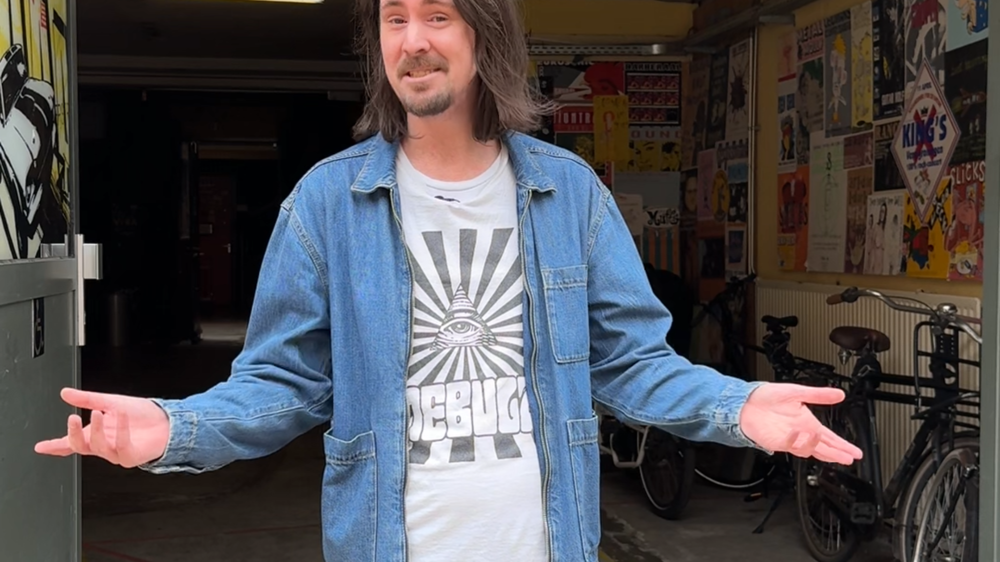 Artiestenbegeleider Mark in de deuropening van de garage van VERA. Hij heeft beide armen naar buiten gedraaid, een open houding, en kijkt vriendelijk naar de camera. Alsof hij je verwelkomd in via de artiesteningang van VERA.