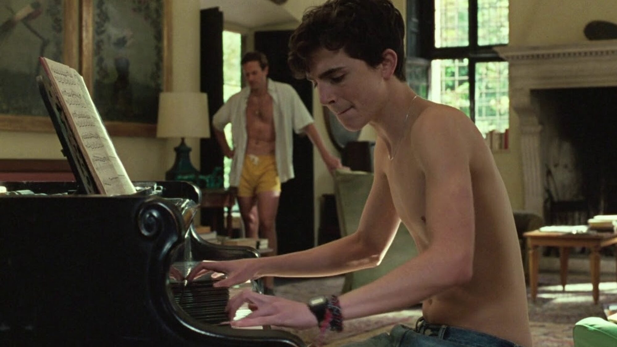 Elio speelt piano in de woonkamer terwijl Oliver staat te luisteren.