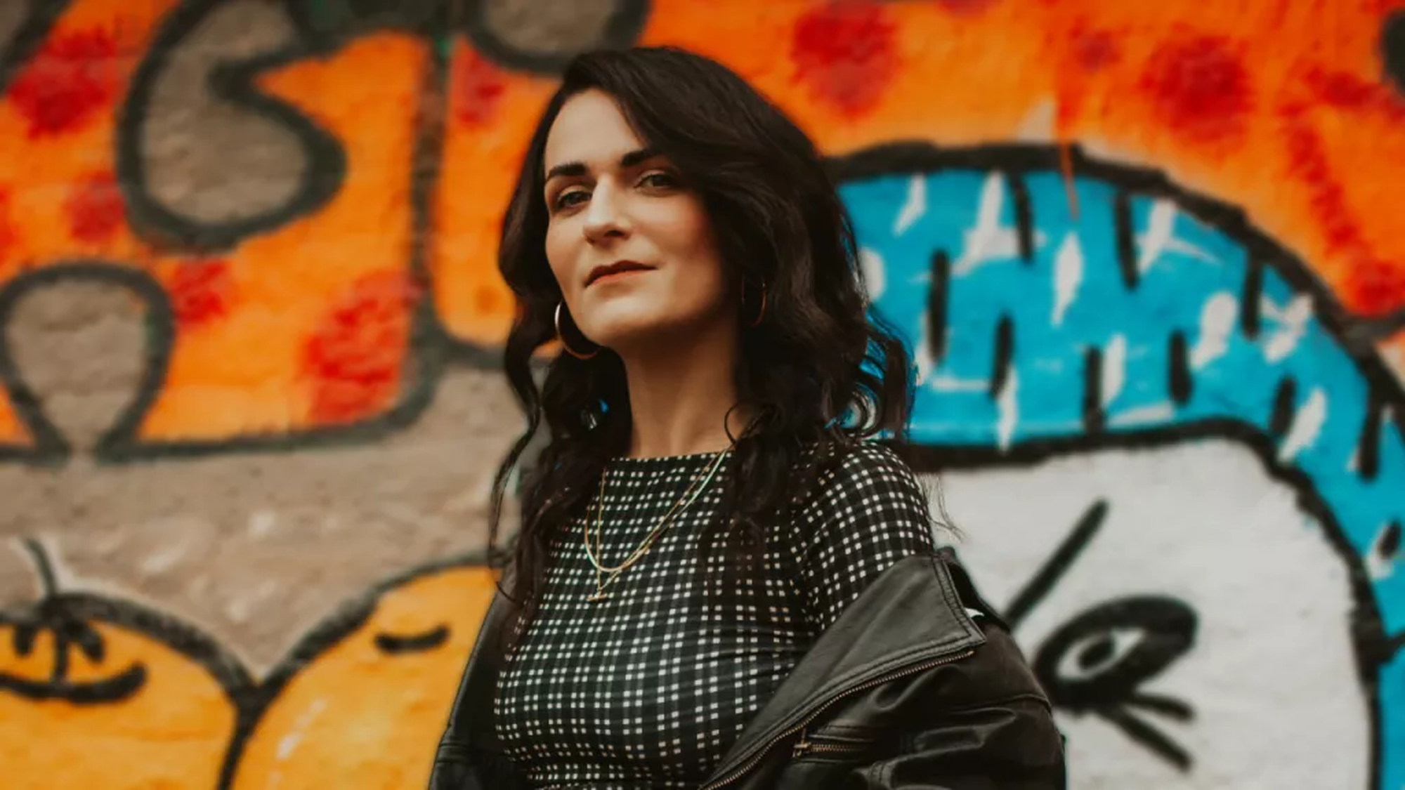 Mevrouw met donkere haren en een nette outfit kijk serieus naar de camera met een gekleurd kunstwerk achter zich