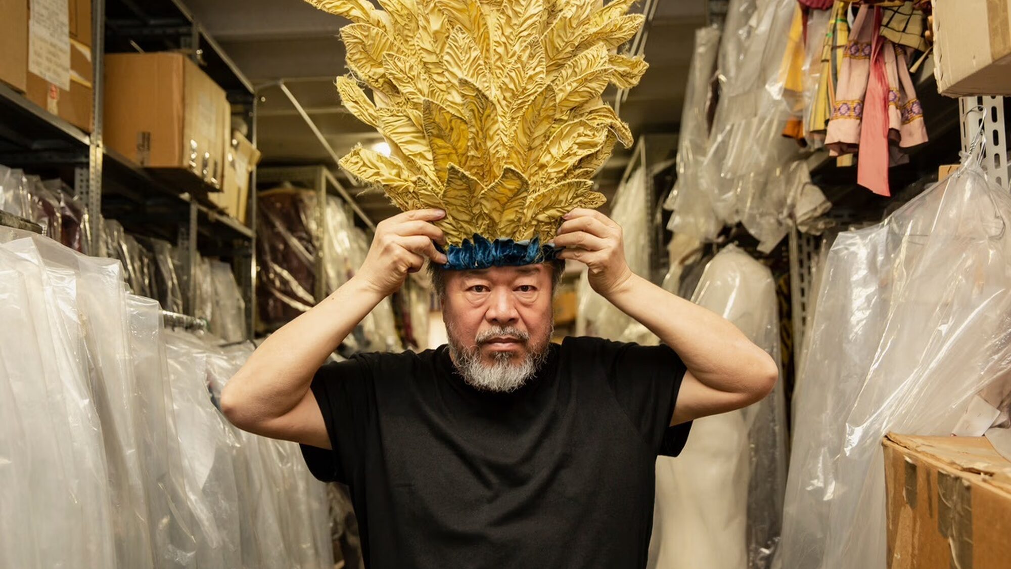 Een stil uit de documentaire waar dit artikel over gaat. we zien ai weiwei, de regisseur, met een imposante gouden tooi op zijn hoofd. Het is een wat oudere man met een grijze baard. Hij zit in een soort kledinghok, met allemaal hangers links en rechts van hem.