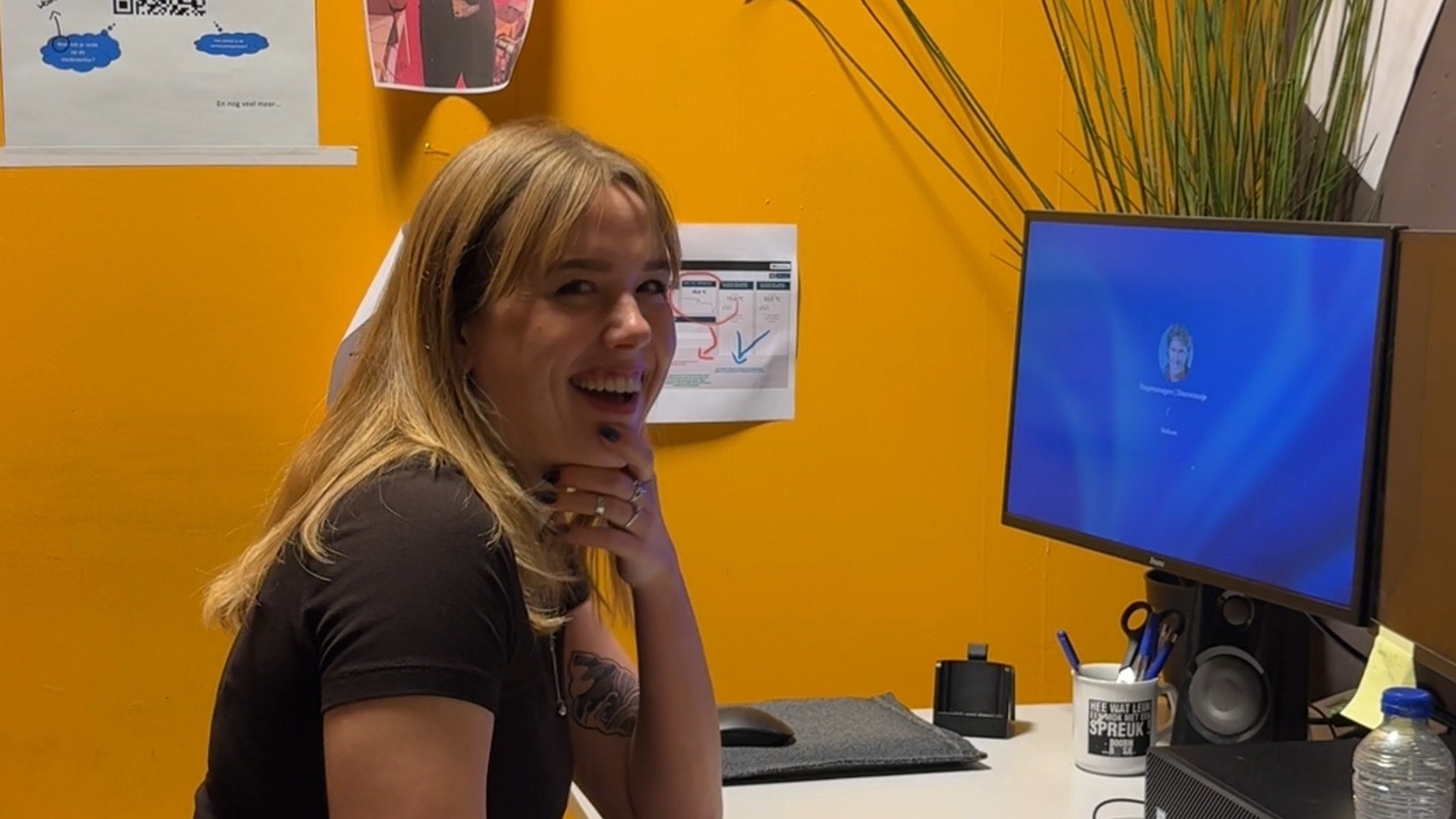 Susanne achter de computer. Ze kijkt lachend naar de camera. De muur is geel en er staat een plant op het bureau.