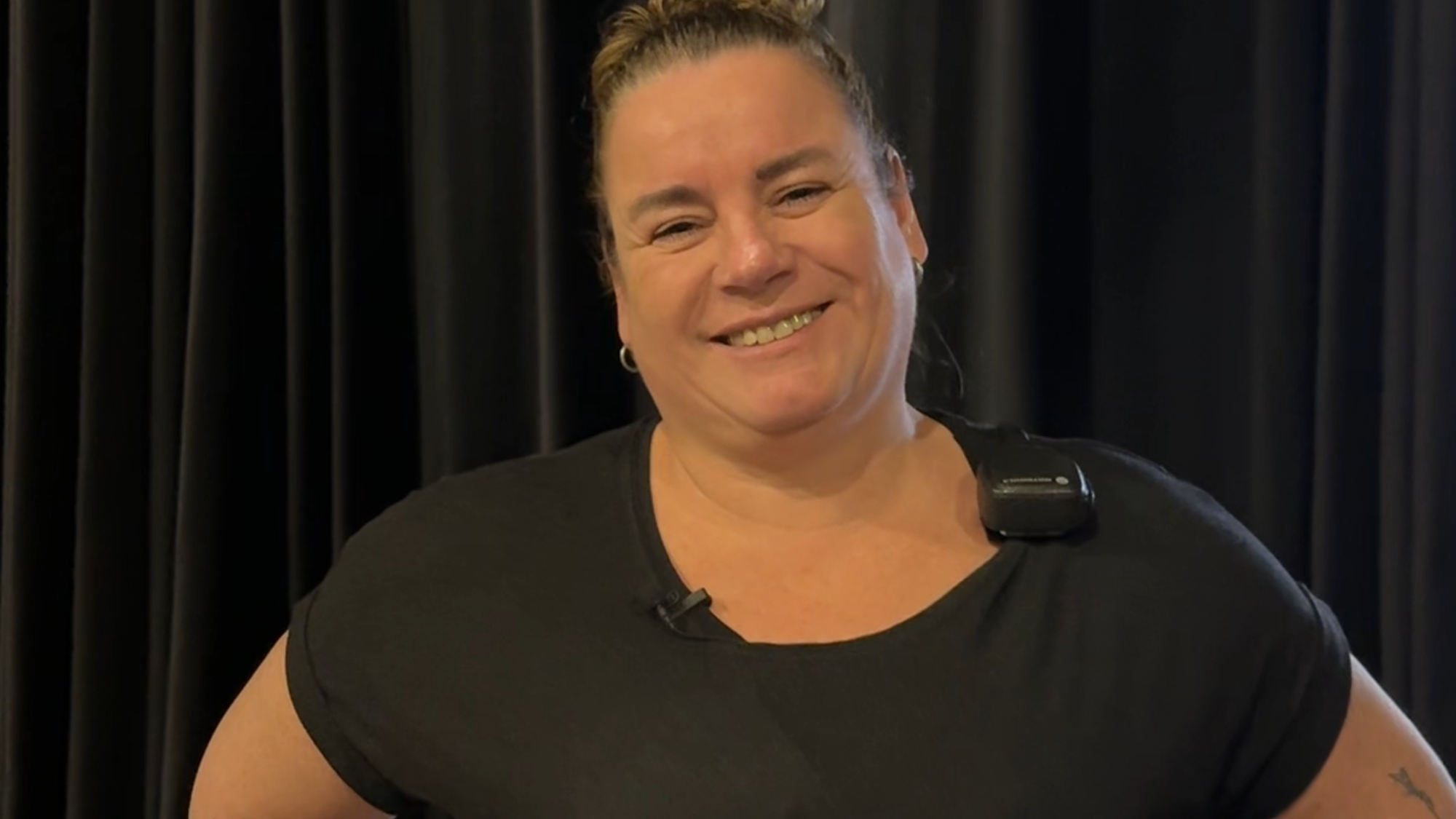 Ilja, die de vragen in dit stuk beantwoordt. Ze staat met haar handen in haar zij en lacht naar de camera. Ze heeft zwarte kleren aan en staat voor een zwart gordijn.