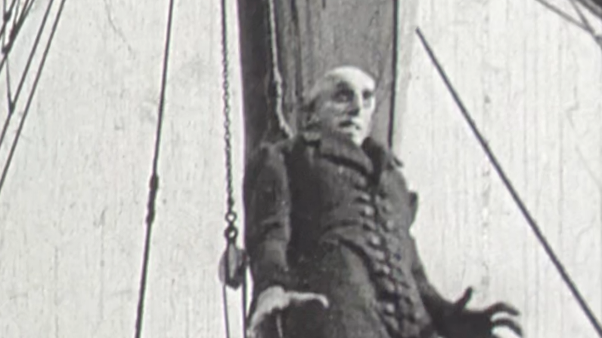 Een stil uit de film Nosferatu. We zien een griezelige man op een schip staan. Hij is erg lang en heeft hele lange vingers.