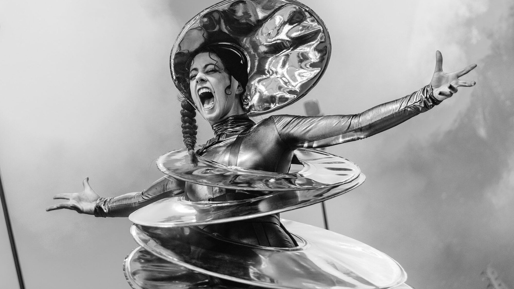 Zwartwitfoto van een vrouw die met haar armen wijd op een podium staat. Haar mond heeft ze wijd open en ze heeft een alienachtig kostuum aan dat bestaat uit een soort schotels. Op haar hoofd draagt ze ook een schotel.