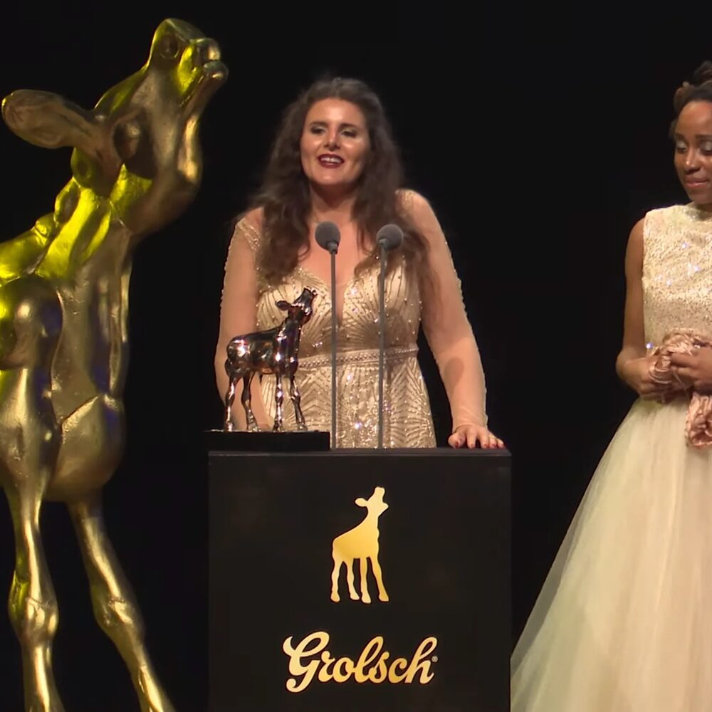 Winnares Gouden Kalf houdt een speech op het podium. Links van haar staat een gigantisch beeld van het Gouden Kalf en rechts staat een vrouw in een witte galajurk.