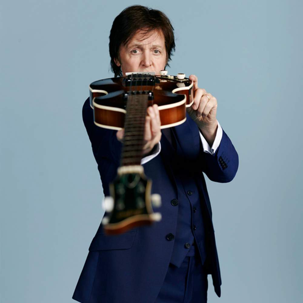 Een studiofoto van Paul McCartney die poseert met zijn basgitaar. Een lichtblauwe achtergrond, Paul met een donkerblauw pak aan en hij houdt de bas met de hals naar de camera toe en kijkt eroverheen.