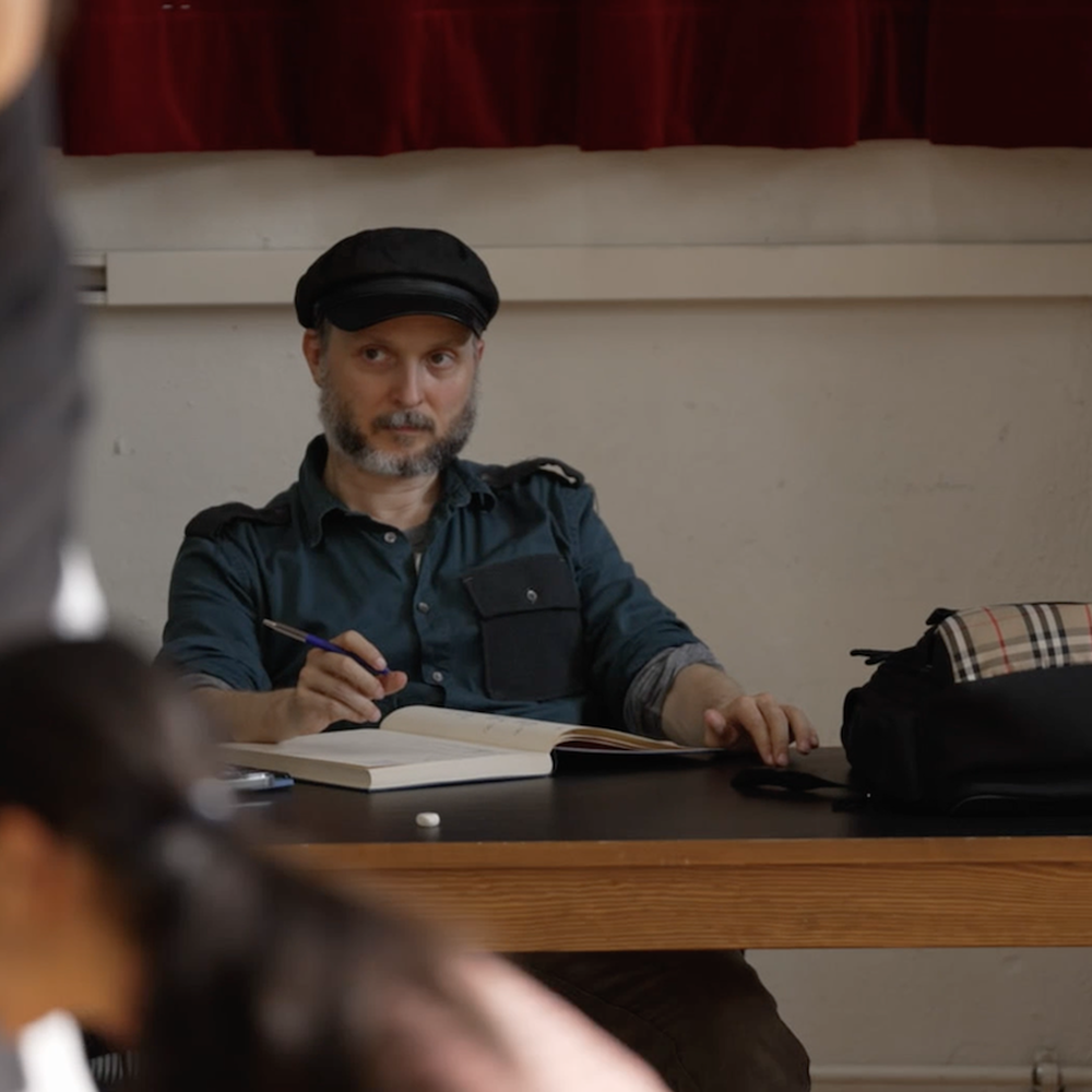 Sidi Larbi Cherkaoui zit met een schrijfboek aan tafel en kijkt naar dansers.