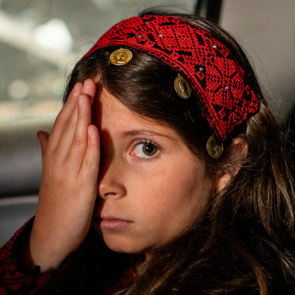 Meisje van vijf houdt een hand over haar linker oog en kijkt de camera in. Ze zit op de achterbank van een auto