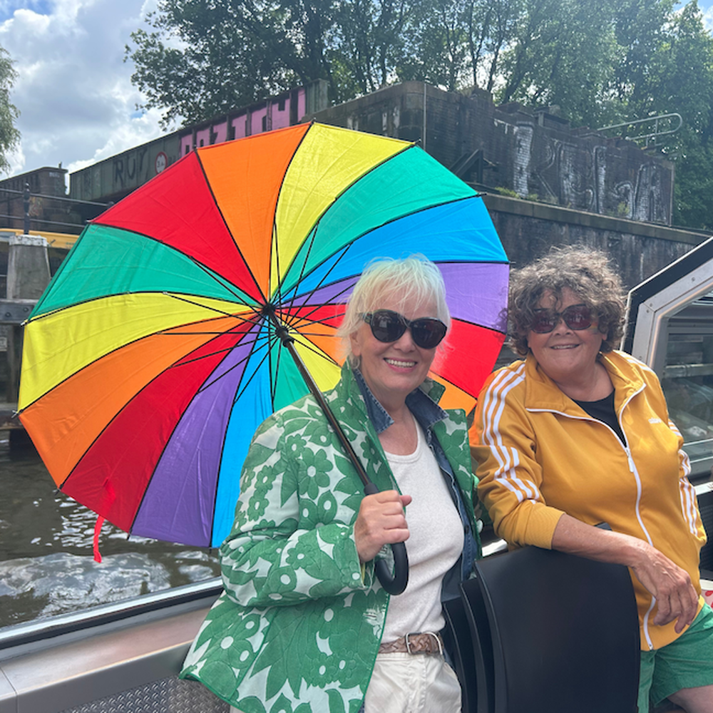 Twee dames op het dek van de boot. Links heeft een mevrouw een een regenboogparaplu vast.