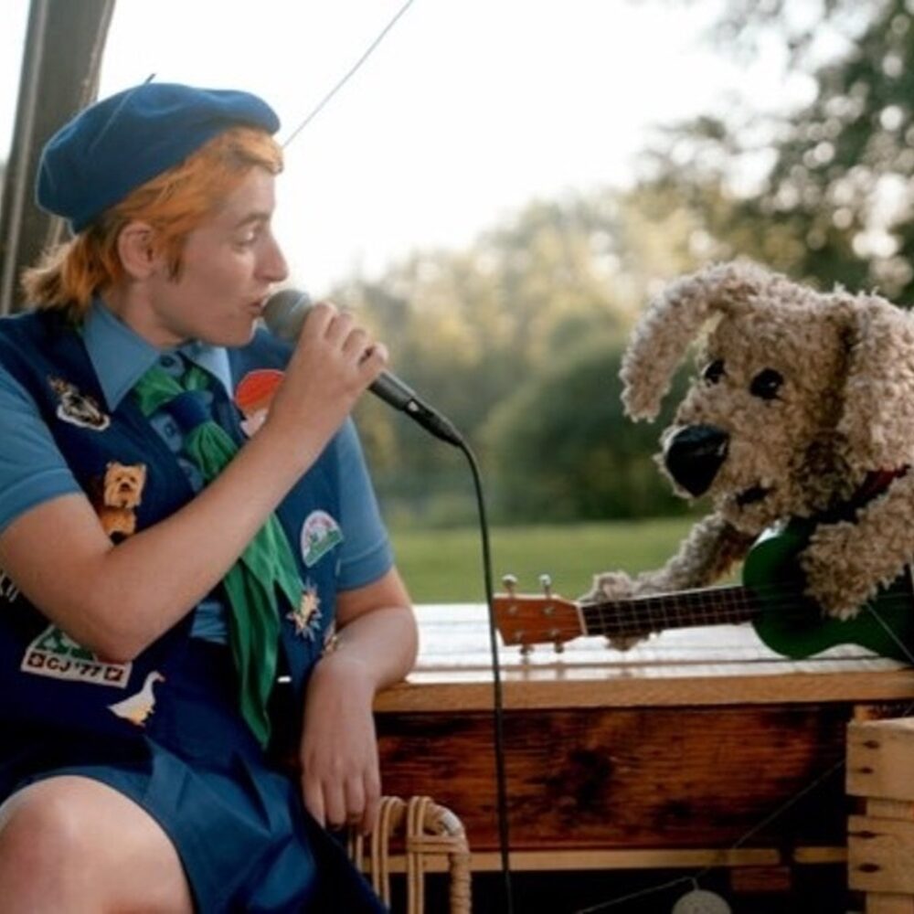 Een foto van Elmer tijdens een concert. Ze heeft een microfoon in haar hand en houdt die bij haar mond. Naast haar zit een pop van een hond met een gitaar in zijn hand. Ze zitten op een bootje.