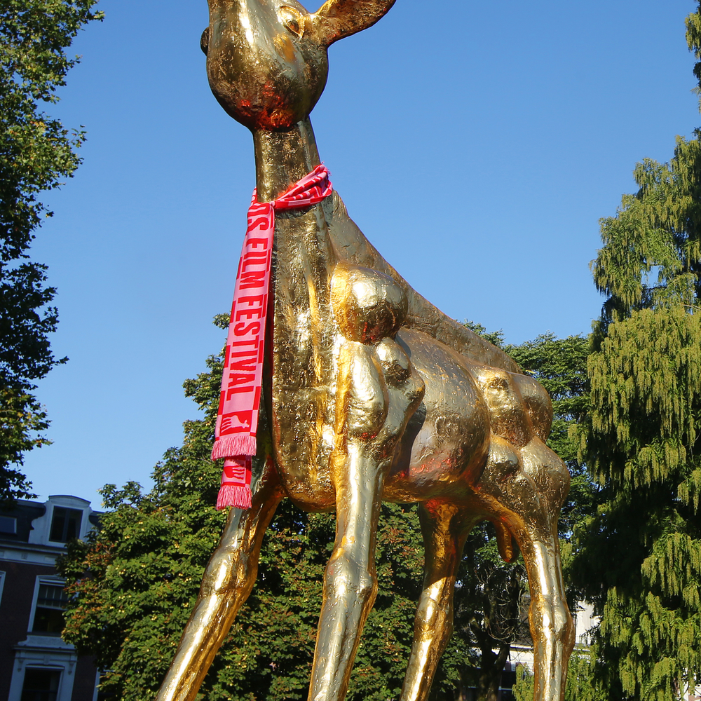 Een foto van een ontzettend groot beeld van een gouden kalf. Het beeld heeft een roze met rode sjaal om. Achter het kalf is een strakblauwe lucht en bomen te zien.