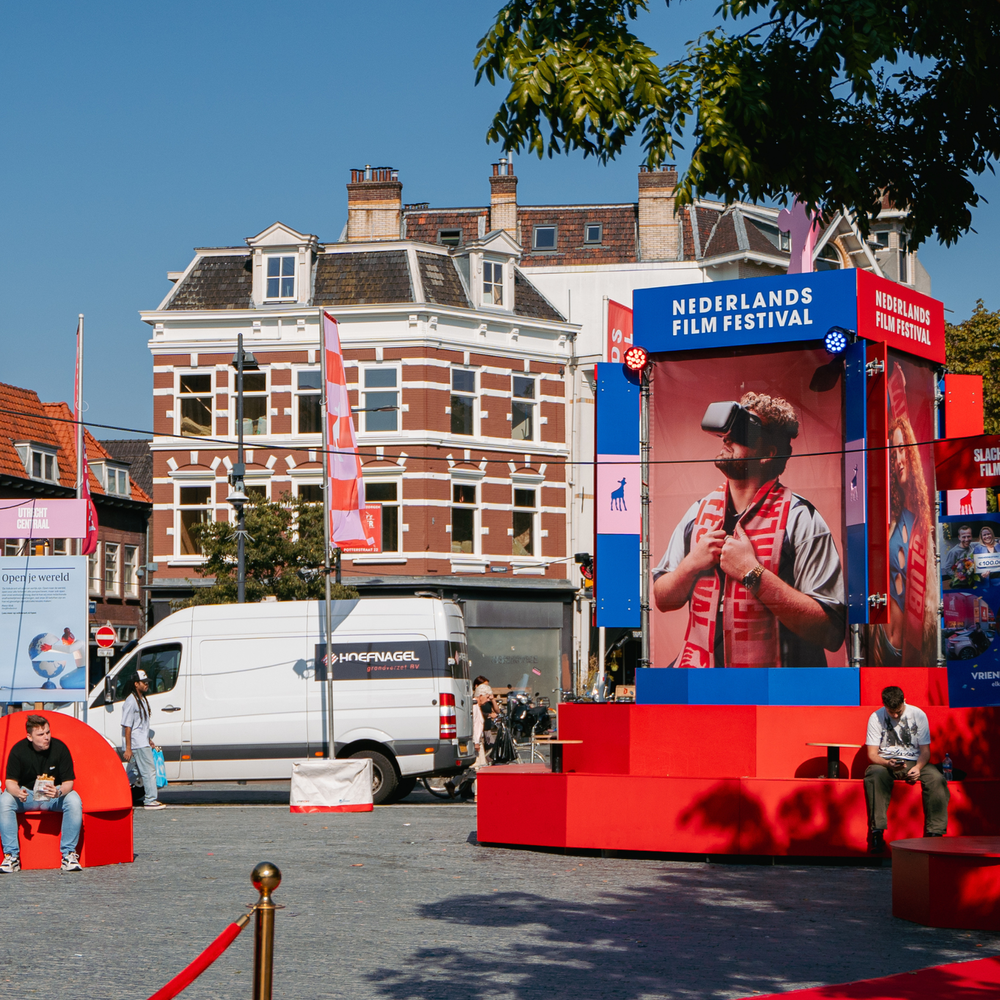 Een foto van het plein Neude in Utrecht tijdens NFF. Je ziet verschillende uitingen van het festival, een informatie stand, vlaggen en een rode loper. Er zitten mensen op bankjes op het plein. Het geheel ziet er feestelijk uit.
