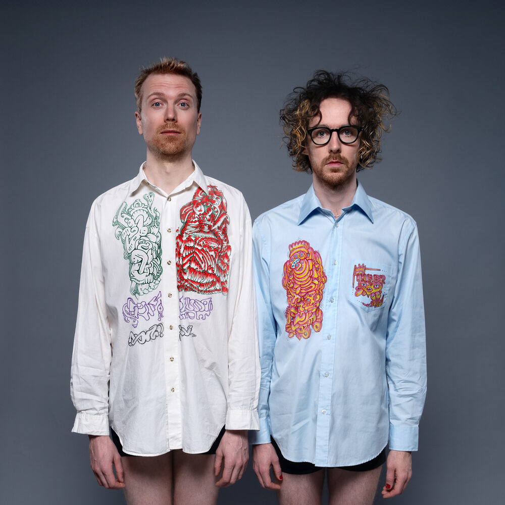 Persfoto van de band Ciao Lucifer. Er staan 2 mannen naast elkaar voor een grijze achtergrond. De linker is blond en de rechter heeft donkere krullen en een bril. Ze dragen beide een lang overhemd met drukke print erop en geen broek.