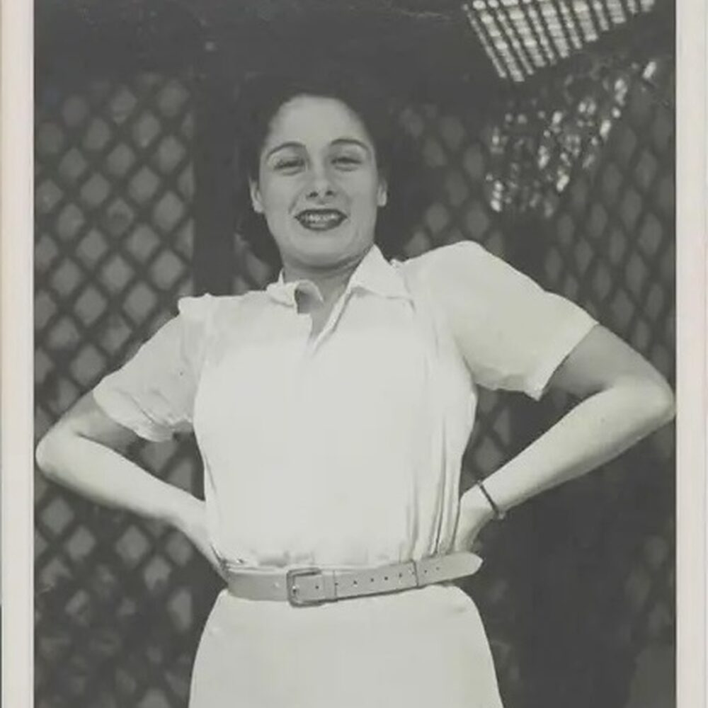 Gaby op een oude foto. Ze kijkt vrolijk in de camera en heeft haar handen in haar zij.