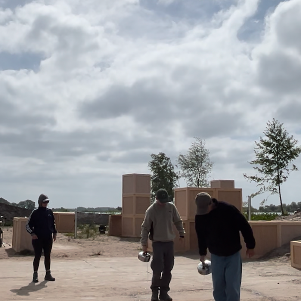 Stil uit het stuk op locatie. De twee hoofdrolspelers gieten water uit een fluitketel op het zand van de zandafgraving.