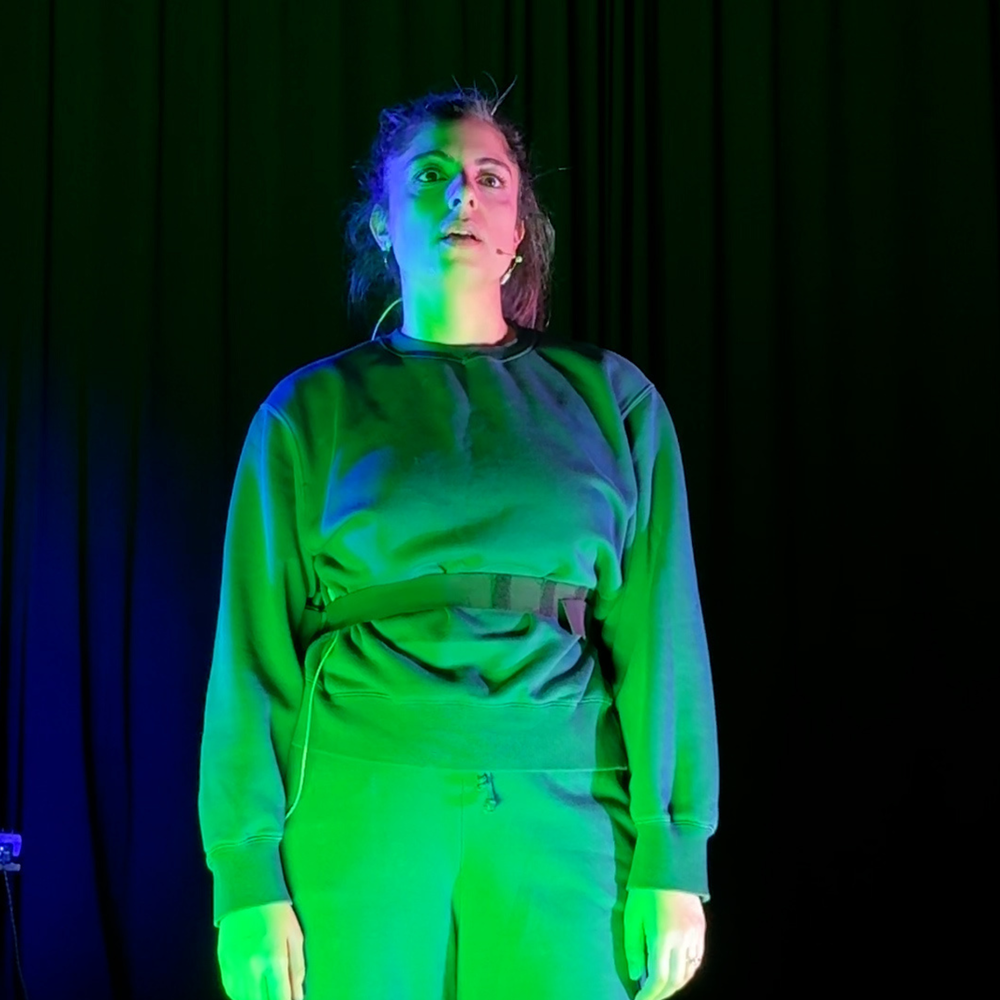 Foto van Yeliz op het toneel. Ze heeft een blauw pak aan en is groen uitgelicht. Ze kijkt serieus.