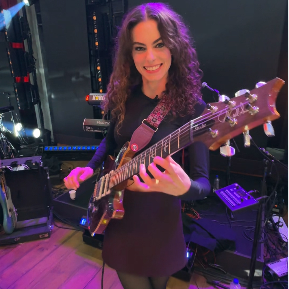 We zien Yvette met haar gitaar in haar handen. Ze staat in een tv studio en kijkt de camera in en lacht. Ze speelt op haar gitaar.