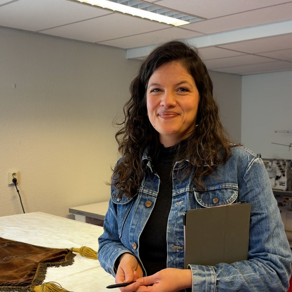 Foto van Lorena in haar atelier. Ze heeft bruin haar loshangend en een spijkerpak aan. Ze heeft een tablet en pen in haar hand en lacht naar de camera.