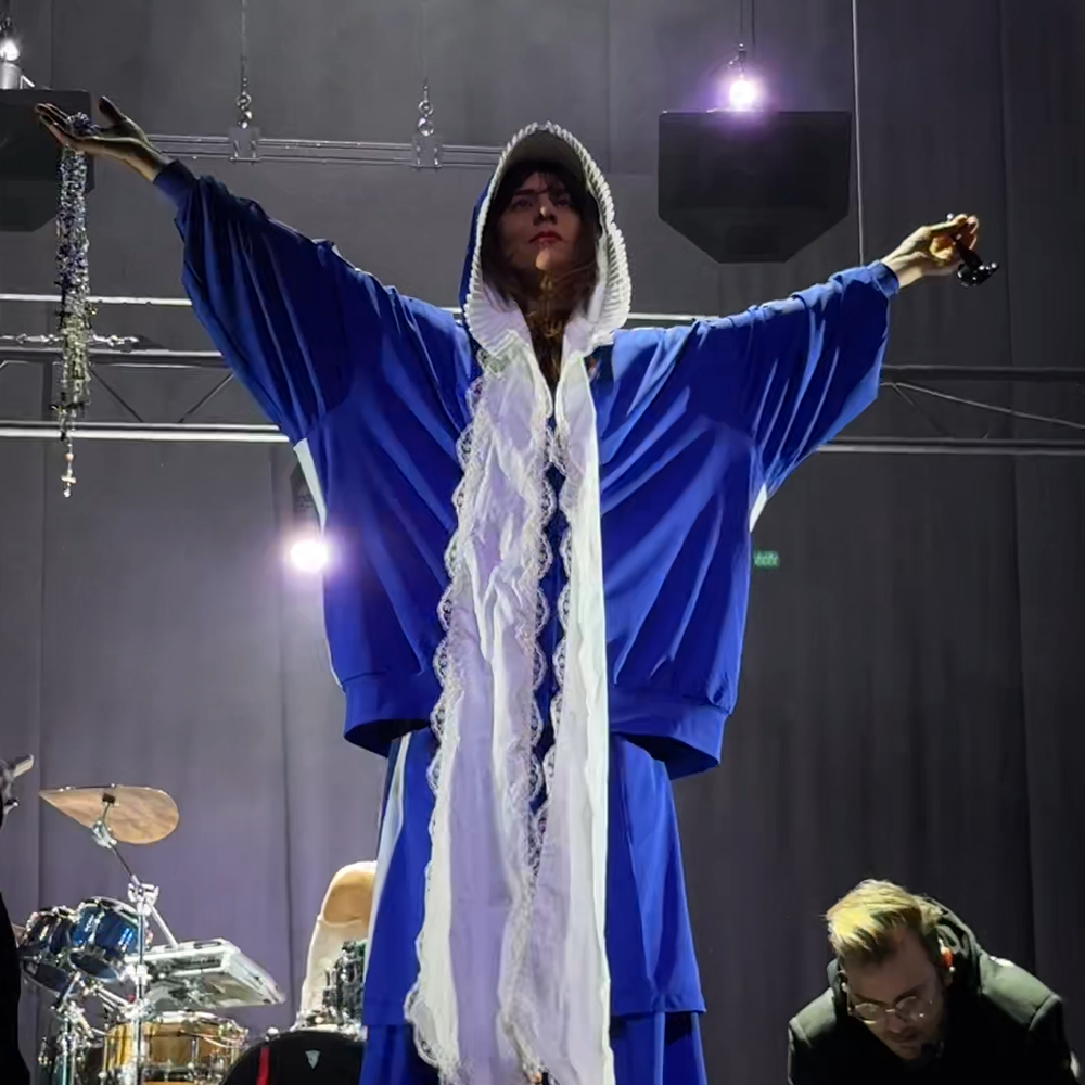 Naomi staat in een blauw met wit pak op een podium met haar armen boven haar hoofd. Ze heeft een zonnebril op en om haar heen staan koorleden.