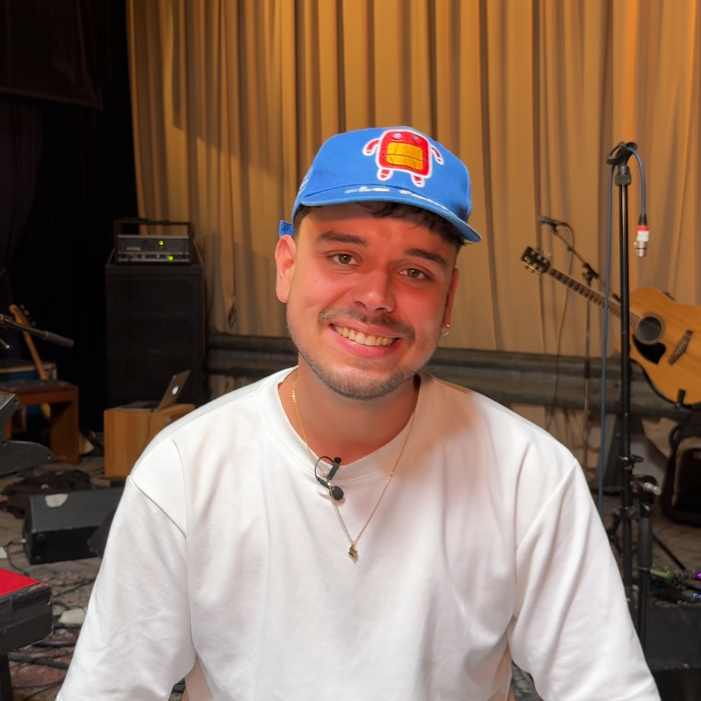 Een foto van de artiest Quique, die getipt wordt in dit stukje tekst. Het is ene jonge jongen met een wit shirt en een pet en hij kijkt lachend de camera in. Hij zit in een repetitieruimte, het is een beetje donker en er hangen gelige gordijnen voor de muur.