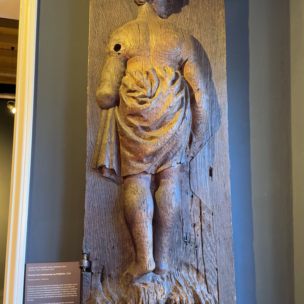 De deur van het pomphuisje. Het is een houten deurtje wat aan een grijze muur hangt. Uit hout gesneden zie je een vrouw op de rug. Ze heeft een doek om haar middel. Ze kijkt naar de rechterkant. Het is houtskleurig.