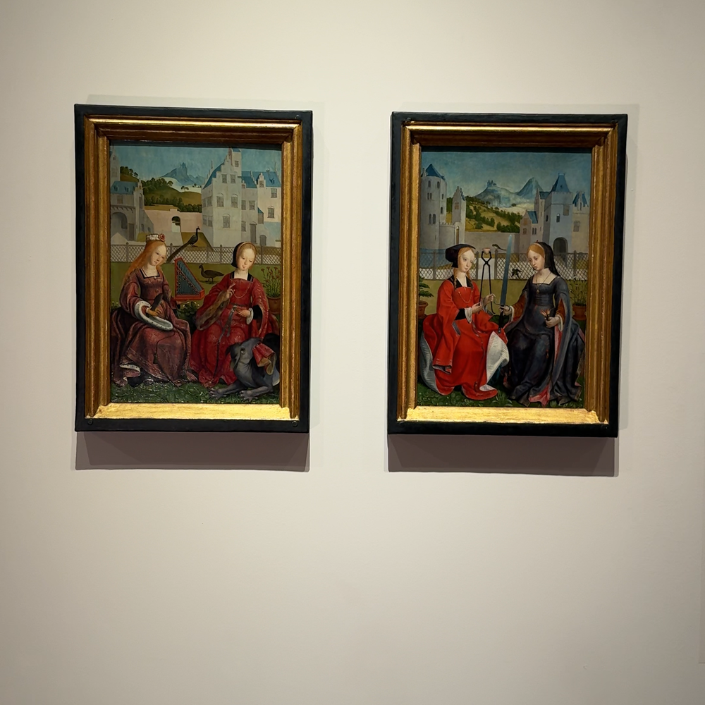 een foto van de twee schilderijtjes die besproken worden in de tekst. Het zijn twee kleine schilderijtjes met vrouwelijke heiligen erop. Ze zitten in gouden lijstjes en hangen naast elkaar. De vrouwen zitten in een soort kloostertuin en hebben allemaal mooie jurken aan met veel goud.