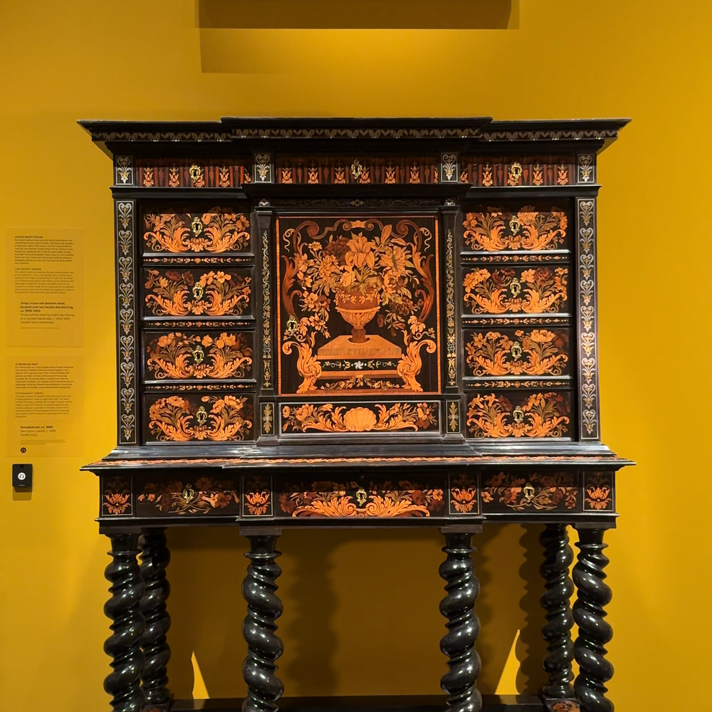 Een foto van de kast die wordt besproken. Het is een ebbenhouten kast, die op poten van ongeveer een meter staat. Er is een bloemenpatroon van hout ingelegd. Hij staat voor een gele muur en erboven hangt een schilderij.