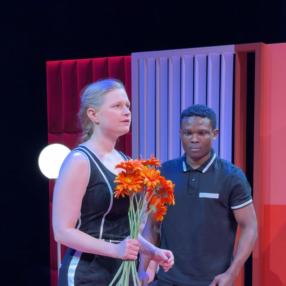 Een stil uit de voorstelling. we zien links een vrouw met oranje bloemen in haar hand en rechts een man die naar de bloemen kijkt. Ze staan stil alsof ze poppen zijn. Het decor waar ze instaan is roze en oranje.