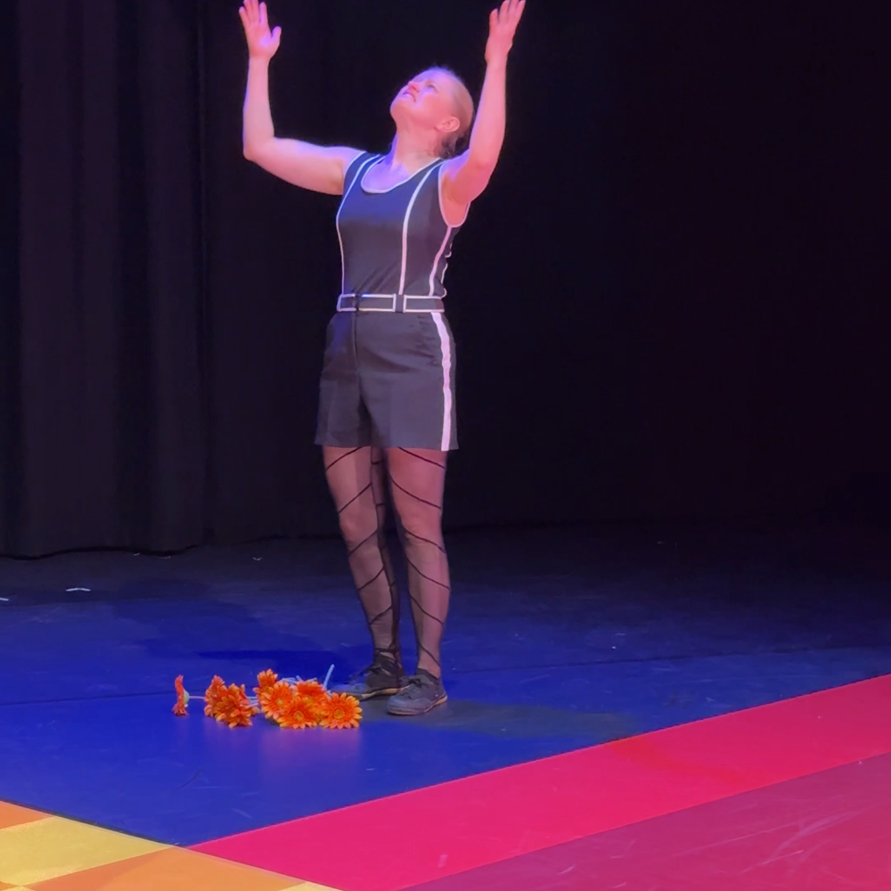 Een stil uit de voorstelling. We zien Roma die de oranje bloemen heeft laten vallen en met haar armen gefrustreerd omhoog beweegt. De voorgrond is een vloer die roze is met een oranje-geel geblokt stuk eroverheen.