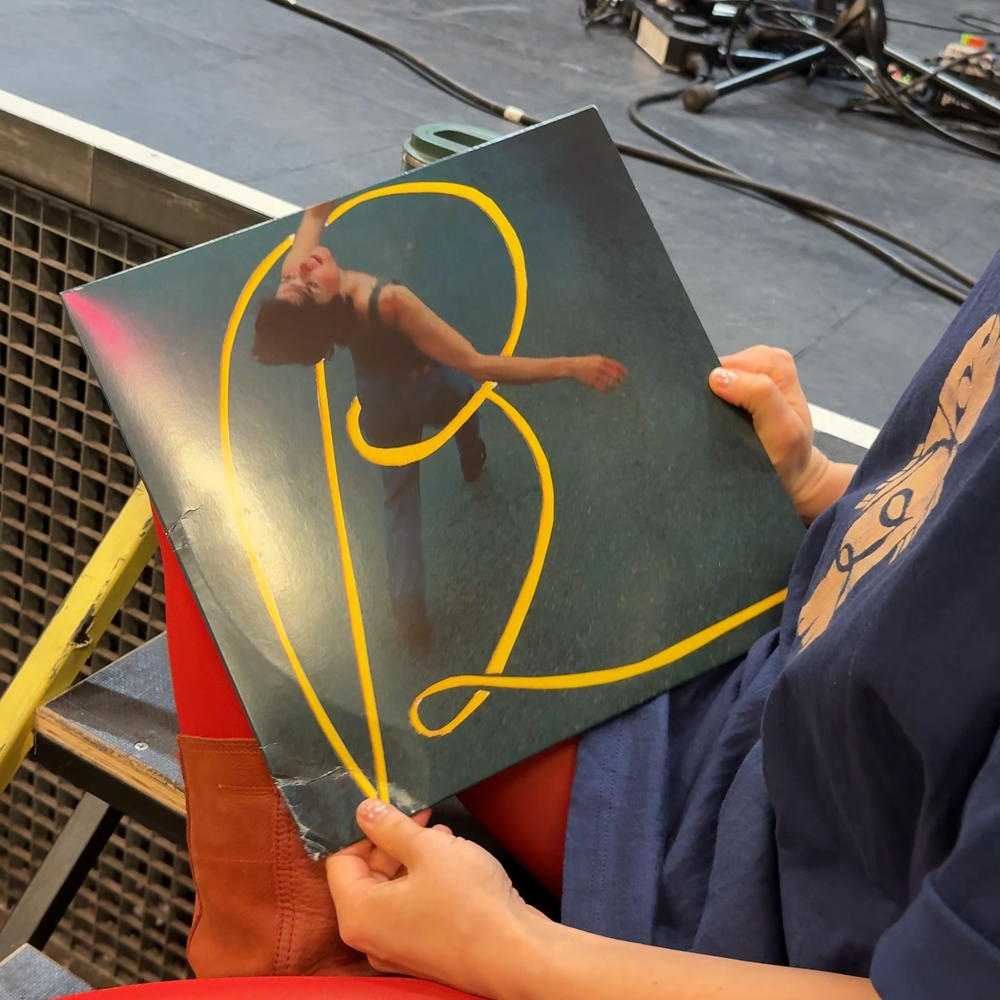 Pitou laat de cover van haar plaat zien. Het is een vinyl plaat met een geel lint erop wat de letters P 2 vormt. Pitou zit op een trapje bij het podium.