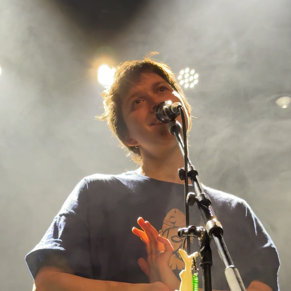 Een foto van Pitou tijdens de repetities. Ze staat op een podium, achter een microfoon en zingt. Ze heeft haar handen tegen elkaar aan. Aan de microfoonstandaard hangt een sleutelhanger met P2 erop.