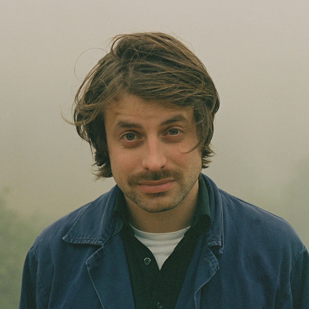 Een portretfoto van Joost Oomen. Een jongeman met wilderig haar, buiten gefotografeerd. Hij heeft een snor en draagt een blauwe jas.