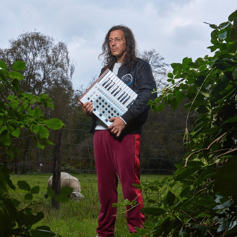 Producer Legowelt met een elektronische piano in zijn armen in een weiland met een schaap op de achtergrond