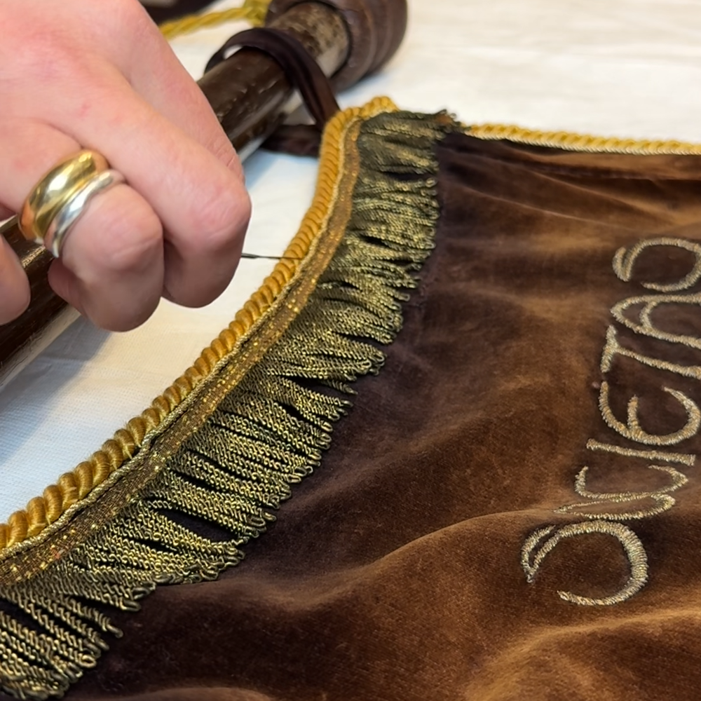Een close-up van het vaandel. Het is bruine fluweel stof. Je ziet Lorena's hand. Er zitten kleine kwastjes aan het vaandel, die zij aan de kant schuift met een metalen instrument.