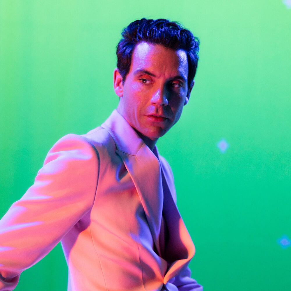 Foto van zanger Mika, in een roze pak voor een groene achtergrond.