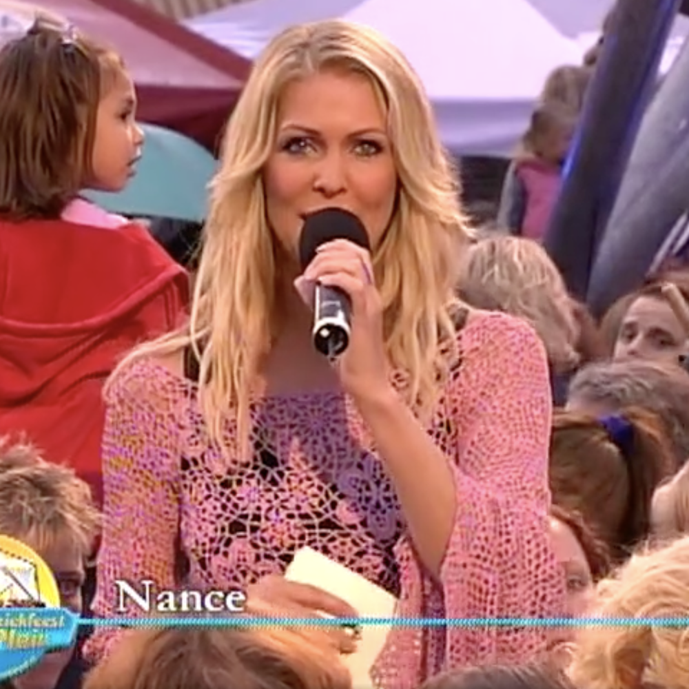 Presentatrice Nance. Ze praat in een microfoon en heeft een roze top aan. Onder in beeld zien we een naambalkje staan met Nance en het tros muziekfeest logo. Ze staat middenin het publiek. Het is een beeld uit de uitzending van 2005 en dat is ook te zien aan het logo en de beeldkwaliteit.