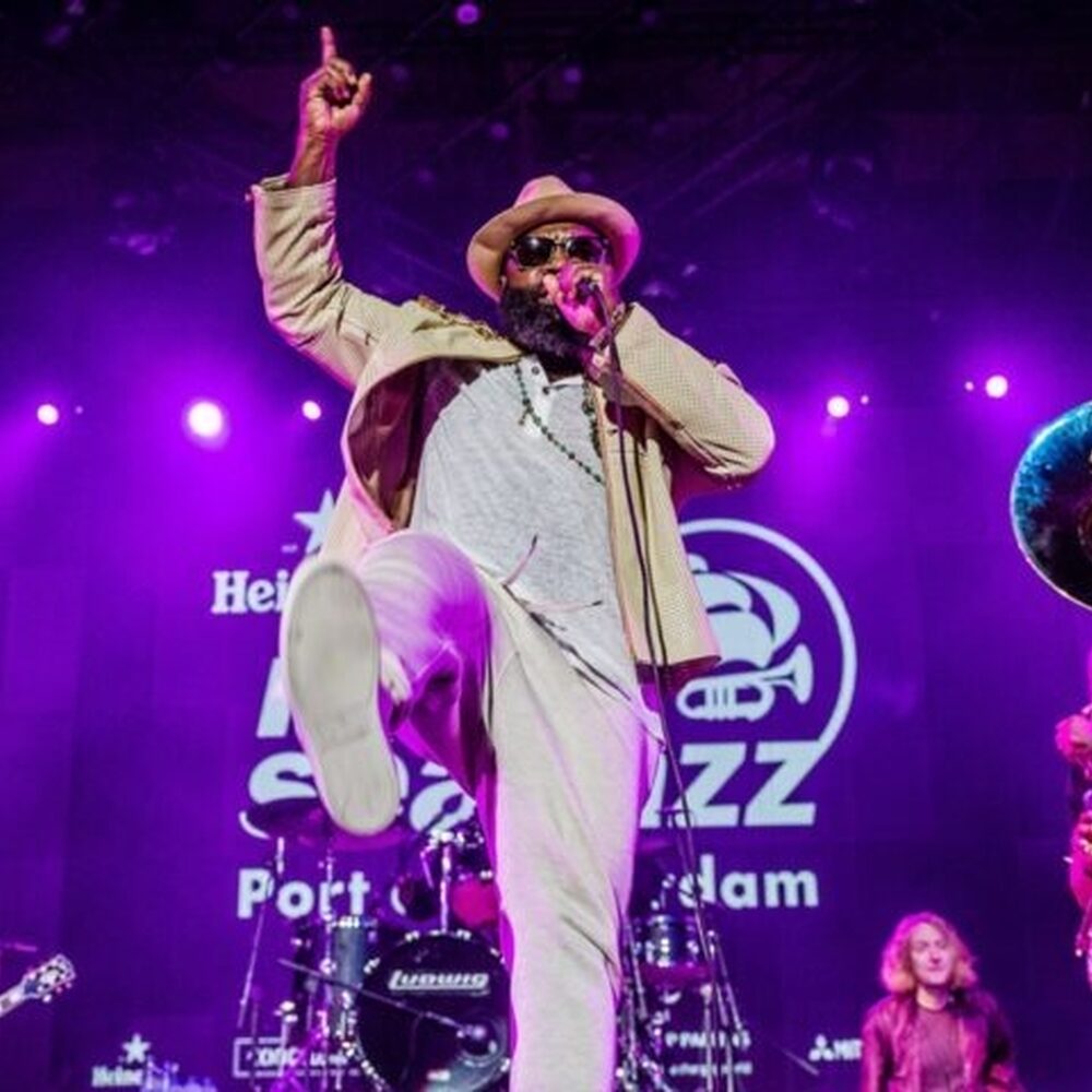 Een zanger danst en zingt op het podium van North Sea Jazz. Achter hem staan muzikanten en het podium is paars verlicht.