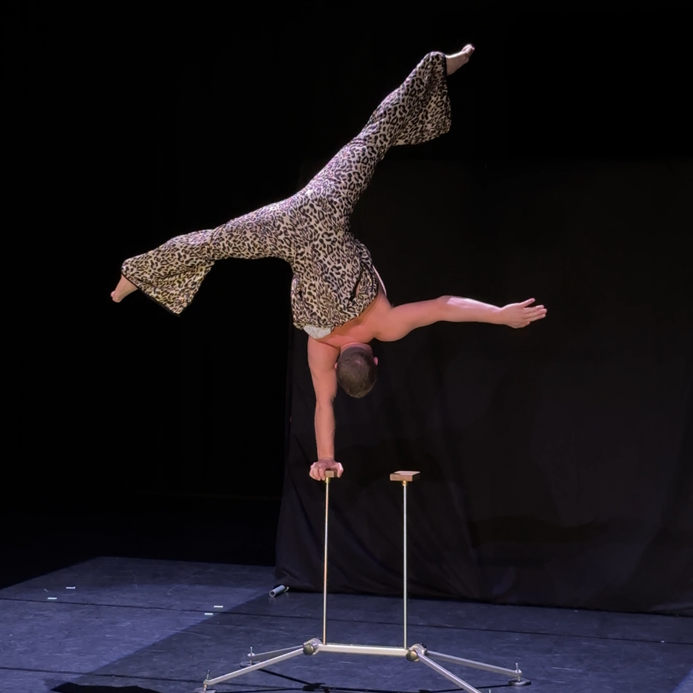 Een foot van Nick die een handstandmaakt op een speciale houder daarvoor. Het is een volledig zwarte achtergrond, zwarte vloer en Nick draagt een tijgerprintpak.