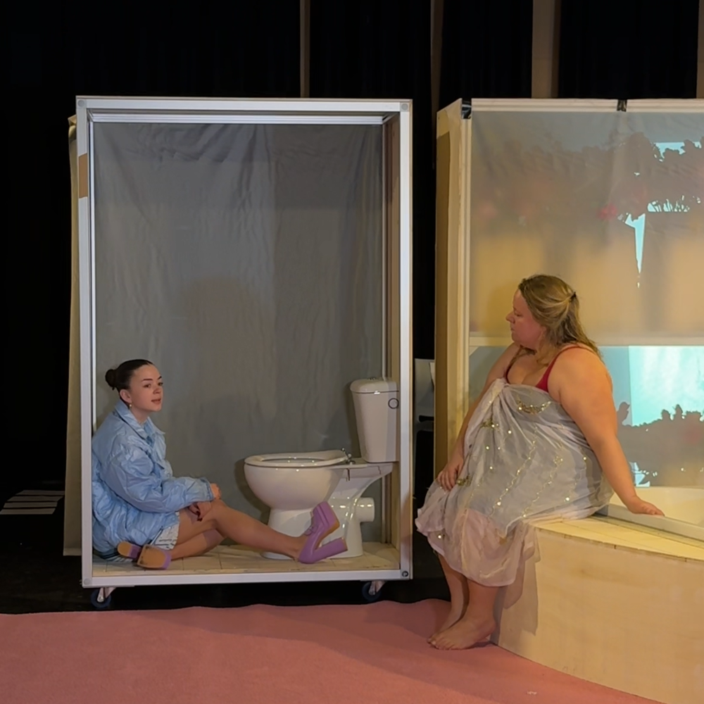 De twee jongere actrices uit het stuk zitten samen in het decor. De ene zit naast een wc en de ander op de badrand.