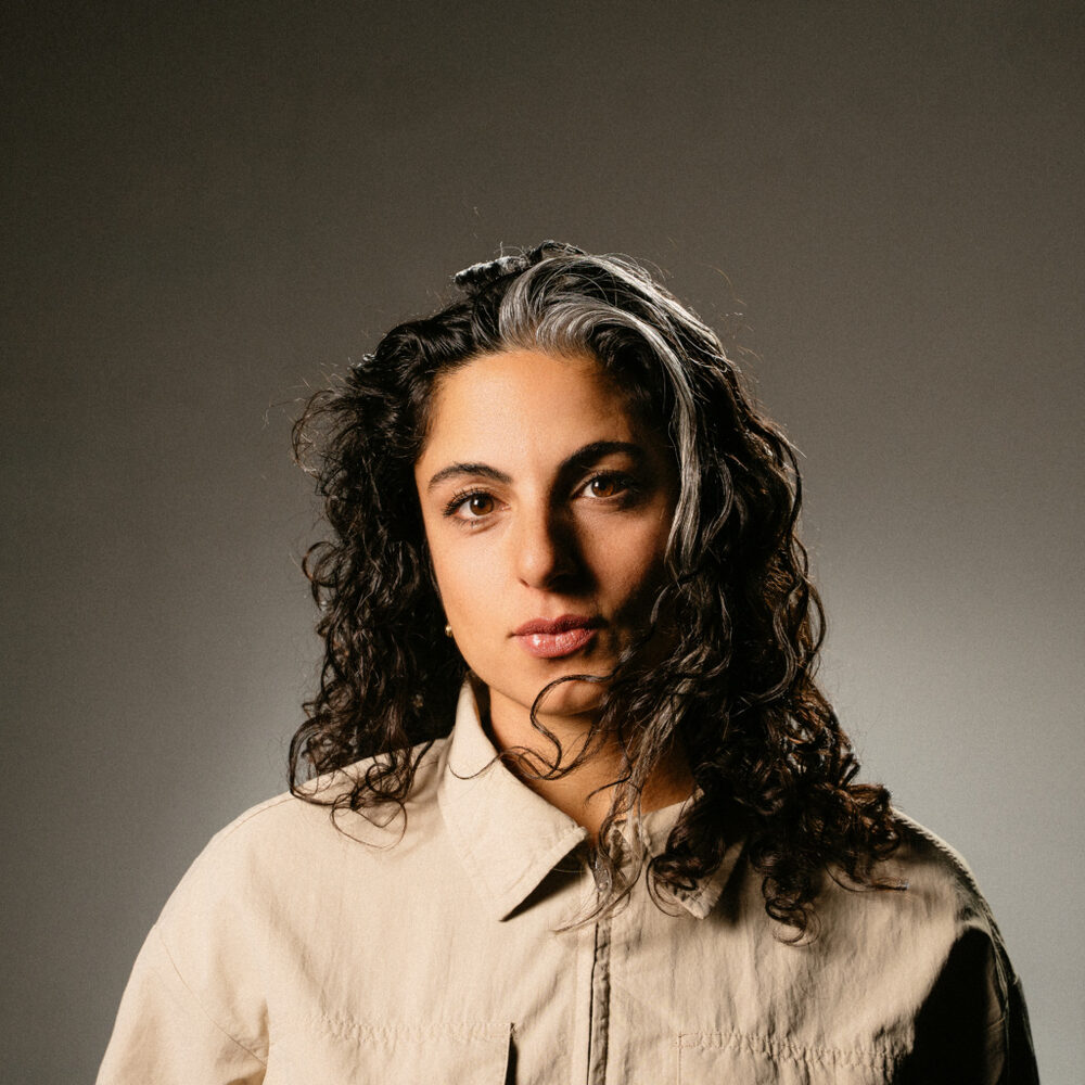 Een foto van Yeliz, de actrice over wie dit stuk gaat. Ze kijkt te camera in en staat voor een bruine achtergrond. Haar zwarte krullen hangen los langs haar hoofd en ze draagt een lichtbruine blouse.