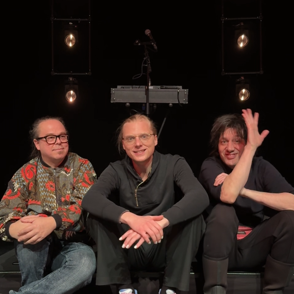 De drie leden van de band Prins S en de Geit zitten op een klein podiumpje en kijken naar de camera. Ze lachen allemaal vriendelijk en de meest rechter zwaait.